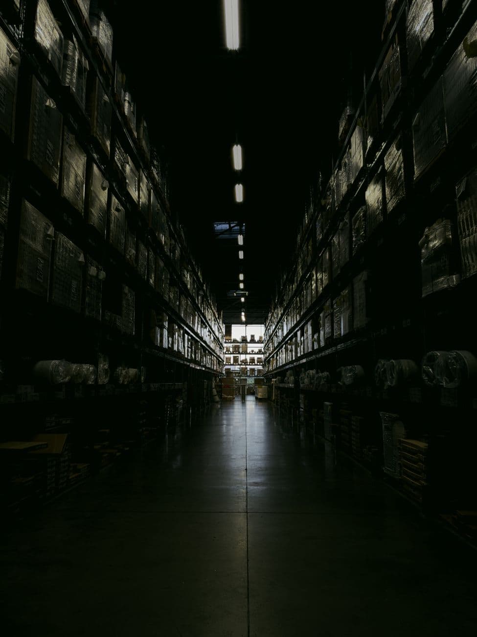 A dim warehouse aisle lined with tall stocked shelves, receding to a far doorway.